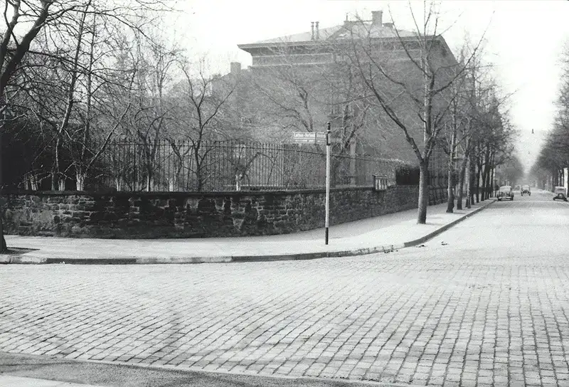 Schwarz-weiß Bild einer Straßenecke. Im Vordergrund die Straße mit Kopfsteinpflaster, dahinter eine Mauer mit Zaun und einige große Häuser.