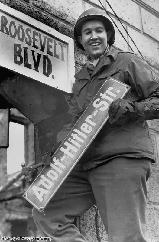 Altes schwarz-weiß Foto eines amerikanischen Soldaten, der das Straßenschild "Adolf-Hitler-Straße" gegen das Schild "Roosevelt Blvd." austauscht. Er lächelt dabei froh nach rechts unten.