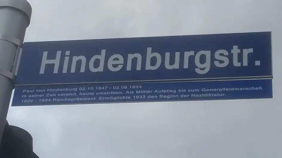 Straßenschild "Hindenburgstraße" mit Zusatztafel vor grauem Himmel. Auf der Zusatztafel unterhalb des Straßenschildes steht: "Paul von Hindenburg 02.10.1847 - 02.08.1934. In seiner Zeit verehrt, heute umstritten. Als Militär Aufstieg bis zum Generalfeldmarschall. 1922 - 1934 Reichspräsident. Ermöglichte 1933 den Beginn der Nazidiktatur."