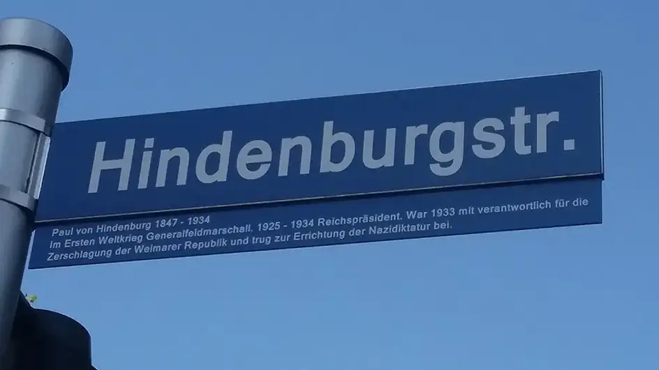 Straßenschild "Hindenburgstraße" mit Zusatztafel vor blauem Himmel. Auf der Zusatztafel unterhalb des Straßenschildes steht: "Paul von Hindenburg 02.10.1847 - 02.08.1934 Im Ersten Weltkrieg Generalfeldmarschall. 1925 - 1934 Reichspräsident. War 1933 mit verantwortlich für die Zerschlagung der Weimarer Republik und trug zur Errichtung der Nazidiktatur bei."