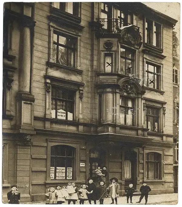 Altes Foto einer Häuserfassade von 1938. Schwarz-Weiß-Fotografie mit leichter Sepia-Tönung. Vor dem Haus steht eine Gruppe Personen, Erwachsene und Kinder.