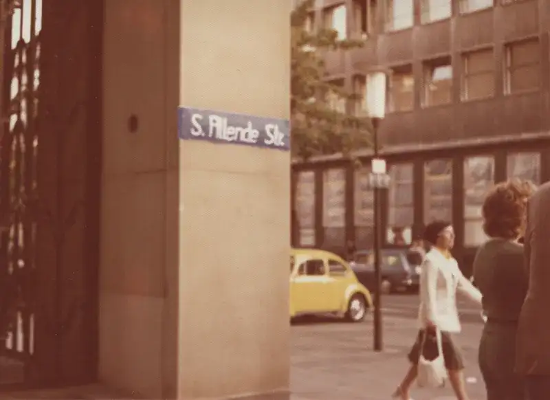 Foto von 1974, zeigt die Gebäude-Ecke mit dem selbst gemalten Straßenschild. Im Hintergrund sind eine Häuserfront, Autos und Personen abgebildet.